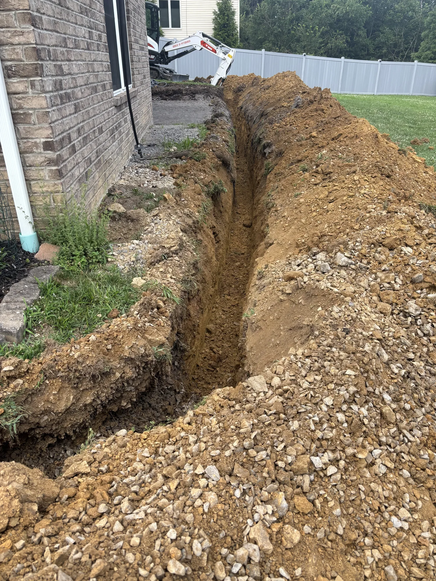 Compact excavator digging a utility trench along a house foundation in the North Hills of Pittsburgh